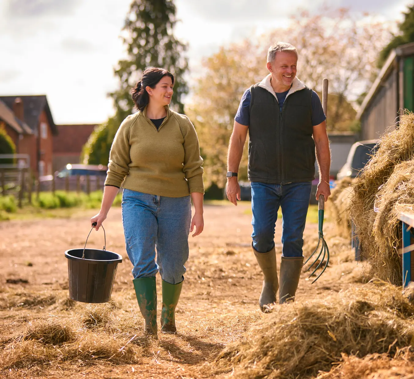Best of british Two farmhands stand in a hay-strewn yard: on the left, a young woman beams with a bright, friendly smile as she grips a black bucket; on the right, a man holds a pitchfork over his shoulder, his brow furrowed and jaw set in focused determination. Behind them, stacked hay bales, a wooden fence and leafy trees complete the rustic scene.